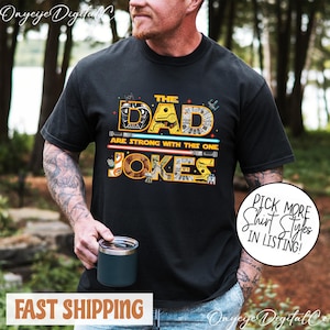Puede incluir: Camiseta negra con un diseño temático de Star Wars. El texto dice "THE DAD ARE STRONG WITH THIS ONE JOKES" en amarillo y naranja. El diseño incluye sables de luz y otros elementos de Star Wars.