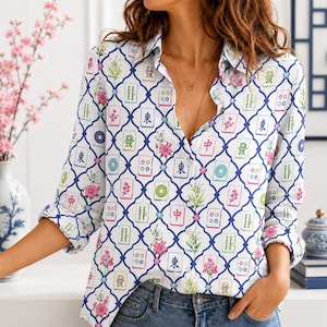 May include: A long-sleeved button-down shirt featuring a white base with a repeating blue lattice pattern. The design includes colorful illustrations of mahjong tiles and pink floral sprigs distributed throughout the fabric. The shirt has a collar.