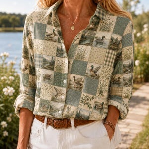 May include: A long-sleeve button-down shirt featuring a patchwork pattern of ducks, forest landscapes, and floral motifs in shades of green, beige, and blue. The shirt is worn tucked into white pants with a brown leather belt and a gold necklace.