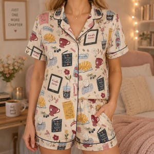 May include: A short-sleeve button-down pajama set in a cream color featuring a repeating pattern of books, e-readers with the text Kindle Girlie, mugs, cats, and flowers. The set includes a collared shirt and matching shorts with dark piping.