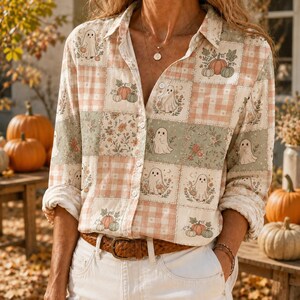 May include: A long-sleeve button-down shirt featuring a patchwork pattern of light orange gingham, sage green, and cream squares. The print includes illustrations of white ghosts, orange and green pumpkins, and small floral arrangements throughout.