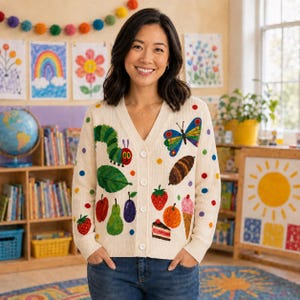 May include: A cream-colored V-neck cardigan featuring a colorful knit pattern of a caterpillar, a butterfly, a cocoon, and various food items including fruit, a slice of cake, and an ice cream cone, accented with small multicolored polka dots.
