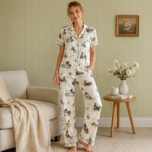 May include: A two-piece pajama set featuring a short-sleeved button-down shirt and matching long pants. The fabric is off-white and decorated with a repeating pattern of mallard ducks in natural colors, accented with green piping along the collar.