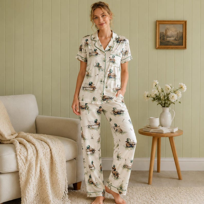 May include: A two-piece pajama set featuring a short-sleeved button-down shirt and matching long pants. The fabric is off-white and decorated with a repeating pattern of mallard ducks in natural colors, accented with green piping along the collar.