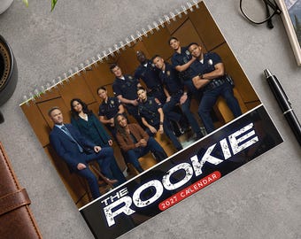 Könnte beinhalten: Ein spiralgebundener Wandkalender für 2027 mit einem Gruppenfoto der Besetzung der Fernsehserie The Rookie. Das Titelblatt zeigt eine Gruppe von Polizeibeamten in Uniform und Berufsbekleidung vor braunem Hintergrund mit dem Titel.