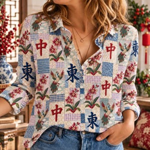 Könnte beinhalten: Ein langärmeliges Hemd mit Knopfleiste und einem Patchwork-Muster aus Blumenmotiven, Phönix-Illustrationen sowie den chinesischen Schriftzeichen 中 und 東 in Rot und Blau. Das Hemd hat einen Kragen und eine helle Grundfarbe.