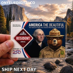 Könnte beinhalten: Eine transparente Plastikkarte mit dem Text "AMERICA THE BEAUTIFUL" und Bildern von George Washington und Smokey Bear. Die Karte enthält auch den Text "RESIDENT ANNUAL" und "AMERICA 250". Der Hintergrund zeigt eine malerische Berglandschaft.