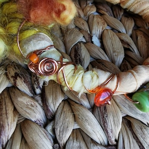 Könnte beinhalten: Ein handgefertigtes Armband mit Kupferdraht, orangefarbenen und grünen Glasperlen und einem beigefarbenen Stoff, der um den Draht gewickelt ist. Das Armband liegt auf einem gewebten braunen Untergrund.