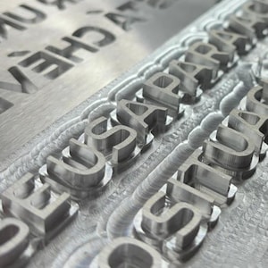 Puede incluir: Primer plano de un sello de metal plateado con las palabras "SEOSAPARATU" y "CONSTITUCION" grabadas en negrita y mayúsculas. El sello tiene una superficie texturizada y probablemente se utiliza para imprimir diseños o texto. El fondo está borroso.