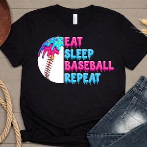 May include: Black t-shirt with a baseball graphic and the text "EAT SLEEP BASEBALL REPEAT". The baseball graphic has colorful dripping icing. A brown cowboy hat and blue jeans are in the background.