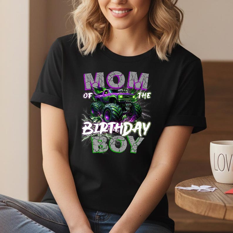 May include: Black t-shirt with a graphic that reads "MOM OF THE BIRTHDAY BOY." The design features a monster truck in green and purple with lightning bolts. The text has a cracked, distressed effect.