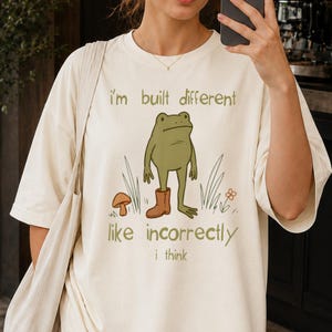 Op de afbeelding: Een crèmekleurig t-shirt met een afbeelding van een groene kikker die één bruine laars draagt. De tekst "i'm built different" staat boven de kikker en "like incorrectly i think" eronder, vergezeld door illustraties van een paddenstoel en gras.