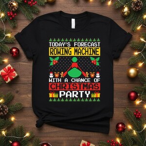 May include: Black t-shirt with a Christmas-themed graphic. The design features the text "TODAY'S FORECAST ROWING MACHINE WITH A CHANCE OF CHRISTMAS PARTY" in a festive, pixelated style, along with rowing machine and holiday elements.