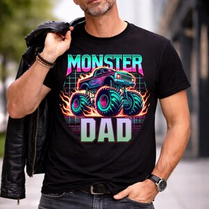 Peut inclure: T-shirt noir avec un graphisme n&eacute;on d'un monster truck et les mots "MONSTER DAD". Le design pr&eacute;sente un camion bleu avec des flammes et de grands pneus turquoise. Le texte est dans une police r&eacute;tro en forme de grille.