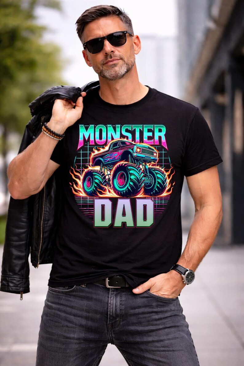 Peut inclure: T-shirt noir avec un graphisme n&eacute;on d'un monster truck et les mots "MONSTER DAD". Le design pr&eacute;sente un camion bleu avec des flammes et de grands pneus turquoise. Le texte est dans une police r&eacute;tro en forme de grille.