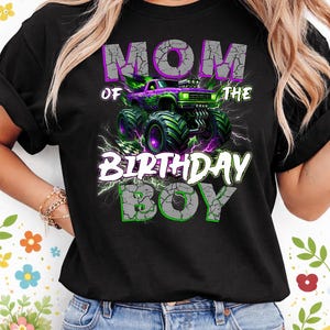 May include: Black t-shirt with a graphic featuring a purple and green monster truck. The text reads "MOM of the BIRTHDAY BOY" in a cracked, distressed font, with lightning bolt accents.