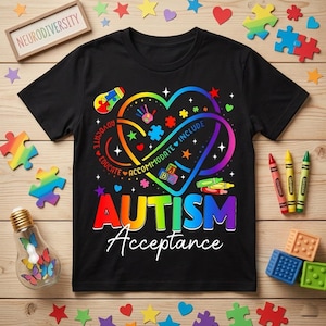 Puede incluir: Camiseta negra con un diseño de corazón colorido y las palabras "AUTISM Acceptance". El corazón incluye piezas de rompecabezas, huellas de manos y las palabras "Advocate, Educate, Accommodate, Include". Un letrero de madera dice "Neurodiversity".