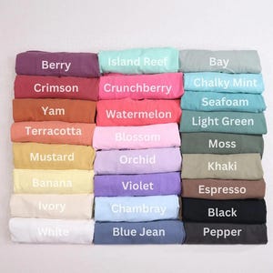 May include: A collection of folded fabric items in various colors, each labeled with a color name. Colors include Berry, Crimson, Yam, Terracotta, Mustard, Banana, Ivory, and White. Other colors are Island Reef, Crunchberry, Watermelon, Blossom, Orchid, Violet, Chambray, and Blue Jean.