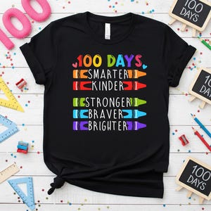 Puede incluir: Camiseta negra con el texto "100 DÍAS" en letras coloridas. Debajo, las palabras "SMARTER, KINDER, STRONGER, BRAVER, BRIGHTER" están escritas con estilo de crayones. La camiseta está decorada con elementos escolares.