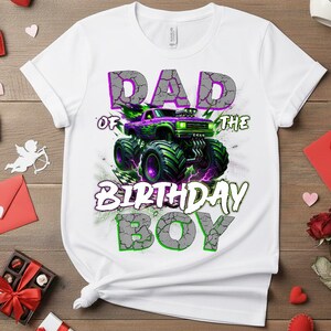 May include: White t-shirt with a graphic of a purple and green monster truck. The text "DAD of THE BIRTHDAY BOY" is printed above and below the truck in a cracked stone font. The shirt is made of a soft material.