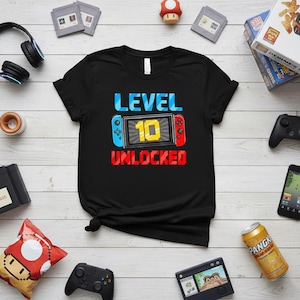 May include: Black t-shirt with the text "LEVEL 10 UNLOCKED" in blue, yellow, and red. The design features a video game controller graphic. The shirt is surrounded by video game accessories and snacks on a light wood-grain surface.