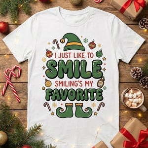 Op de afbeelding: Wit T-shirt met een kerstthema. Het ontwerp bevat een elfenmuts, zuurstokken, ornamenten en de tekst "I just like to SMILE, Smiling's my FAVORITE." Feestelijke kleding voor de feestdagen.