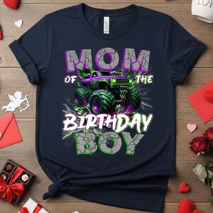 May include: Navy blue t-shirt with a graphic that reads "MOM OF THE BIRTHDAY BOY." The design features a monster truck in purple and green with lightning bolts. The text has a cracked, distressed effect.
