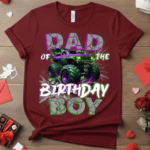 May include: A maroon t-shirt with a graphic featuring a monster truck design. The text reads "DAD OF THE BIRTHDAY BOY" in a distressed font, with a green and purple color scheme. The monster truck has green and purple accents.