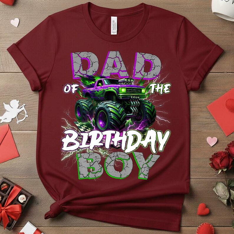 May include: A maroon t-shirt with a graphic featuring a monster truck design. The text reads "DAD OF THE BIRTHDAY BOY" in a distressed font, with a green and purple color scheme. The monster truck has green and purple accents.