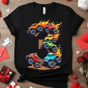 May include: Black t-shirt featuring a colorful graphic of monster trucks arranged in the shape of the number three. The trucks are in orange, red, yellow, green, and blue, with flames and large tires.