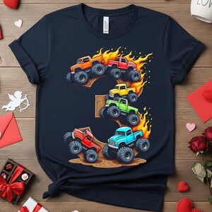 May include: Navy blue t-shirt featuring a colorful graphic of monster trucks in orange, red, yellow, green, and blue, with flames and the number 3. The design is centered on the front of the shirt.