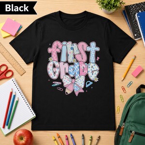 Puede incluir: Una camiseta negra de manga corta con el texto First Grade en letras grandes y estampadas con diseños florales y de cuadros en rosa, azul y blanco. Debajo del texto hay un gráfico de un lazo grande con un lápiz en el centro y estrellas.