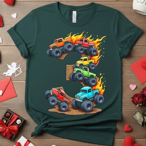 May include: Dark green t-shirt featuring a colorful graphic of monster trucks forming the number 3, with flames. The trucks are in orange, red, yellow, green, and blue. The shirt is made of a soft material.