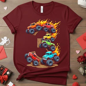 May include: Burgundy t-shirt featuring a colorful graphic of monster trucks in orange, red, yellow, green, and blue, with flames. The trucks are arranged in the shape of the number 3. The shirt is made of a soft material.