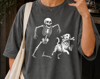 Puede incluir: Una camiseta gris carbón con un estampado gráfico blanco de un esqueleto corriendo y un perro que lleva un hueso en la boca. Al esqueleto parece faltarle un hueso de la pierna, que aparece representado en el suelo junto al perro.