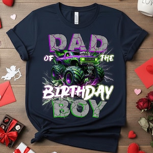 May include: Navy blue t-shirt with a graphic of a monster truck in purple and green. The text reads "DAD OF THE BIRTHDAY BOY" in a distressed font with lightning bolt accents. A fun design for a birthday.