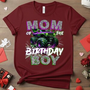 May include: A maroon t-shirt featuring a graphic of a monster truck with the text "MOM OF THE BIRTHDAY BOY". The monster truck is purple and green with large tires. The text is in a cracked, distressed style.
