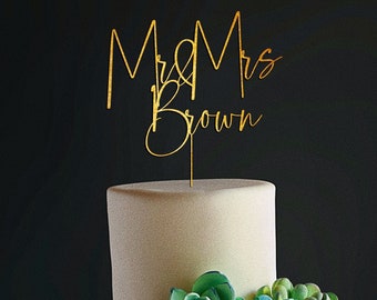 Puede incluir: Pastel de boda de dos niveles decorado con patrones geométricos dorados y suculentas verdes y azules. El adorno de la torta es una escritura dorada que dice "Mr & Mrs Brown".