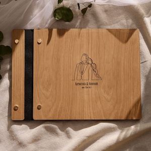 May include: A wooden guest book with a black leather spine and brass accents. The cover is engraved with a line drawing of a couple and the text "Raymond & Hannah, June 17th 2023".