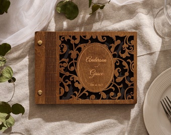 May include: A wooden guest book with a decorative cutout design and the names "Anderson & Grace" and the date "Oct 16, 2021" engraved on the front.