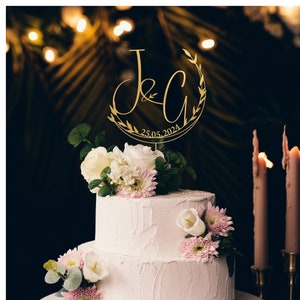 May include: A two-tiered white wedding cake decorated with white and pink flowers. A gold topper with the initials "J & C" and the date "25.05.2024" sits on top of the cake.