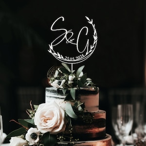 May include: White cake topper with a wreath of leaves and the initials "S&C" with the date "25.05.2024" on a black and white wedding cake.