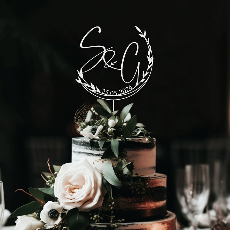 May include: White cake topper with a wreath of leaves and the initials "S&C" with the date "25.05.2024" on a black and white wedding cake.