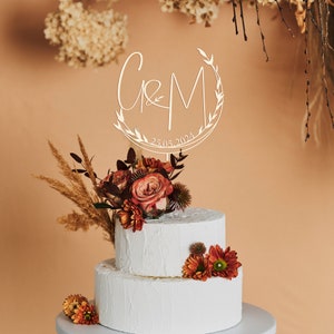 May include: A white two-tiered cake with a wooden cake topper featuring the letters "C & M" in a circle with leaves and the date "25.05.2024". The cake is decorated with a bouquet of red and orange flowers.