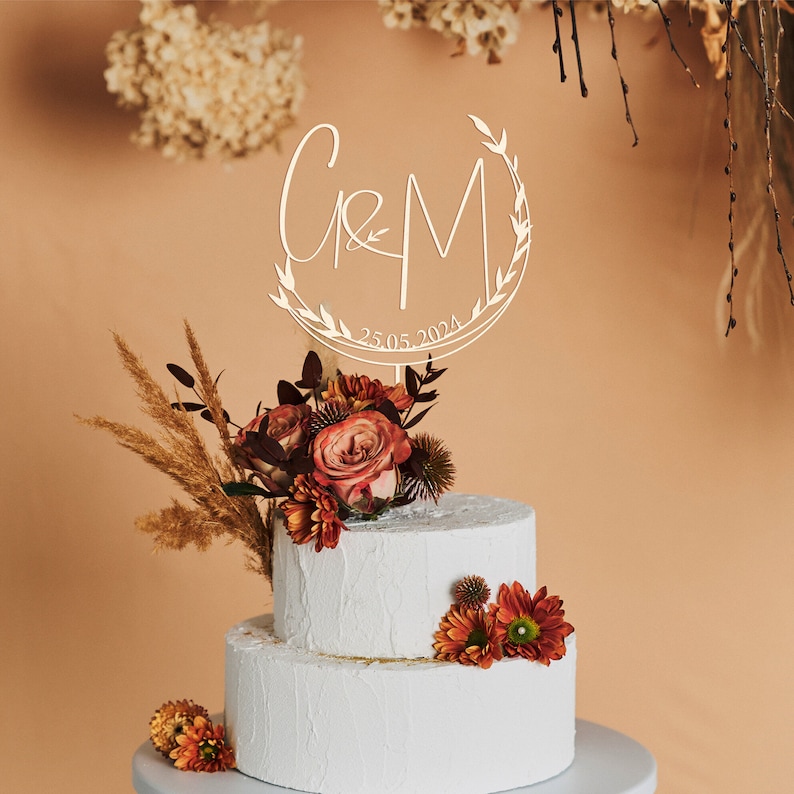 May include: A white two-tiered cake with a wooden cake topper featuring the letters "C & M" in a circle with leaves and the date "25.05.2024". The cake is decorated with a bouquet of red and orange flowers.