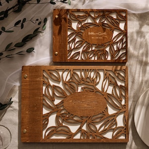 May include: Two wooden photo albums with a leafy design. The top album has the text "William & Gabriella Dec. 1st, 2019" engraved on it. The bottom album has a larger design and the text "William & Gabriella The Ceremony" engraved on it.