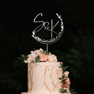 May include: A white wedding cake with a silver cake topper featuring the letters "S&K" in a circle with a leafy design and the date "25.05.2024".