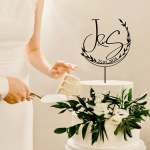 May include: A black wedding cake topper with a floral wreath and the initials "J & S" and the date "25.05.2024".