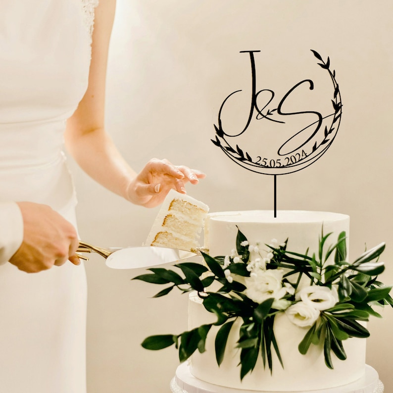 May include: A black wedding cake topper with a floral wreath and the initials "J & S" and the date "25.05.2024".