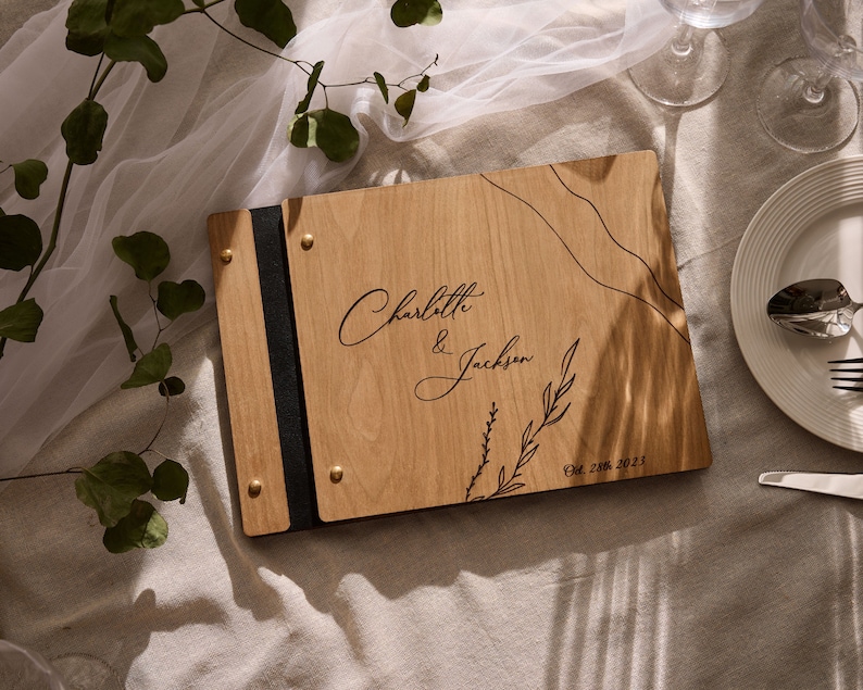 May include: A wooden guest book with a black ribbon closure and gold accents. The cover is engraved with the names "Charlotte & Jackson" and the date "Oct 28th 2023".